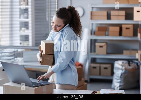 Young busy female worker of post office talking on mobile phone and looking through data on laptop screen by workplace in storage room Stock Photo