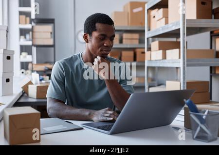 Young serious male manager of storage room using laptop by workplace for checking data about online orders and addresses of receivers Stock Photo
