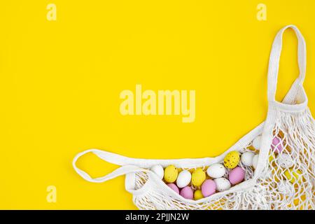 Colored Easter eggs in a mesh bag on a yellow background, flat lay. Stock Photo