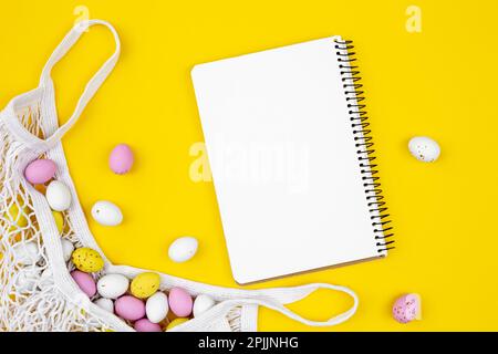 Colored Easter eggs in a mesh bag and a blank notepad on a yellow background. Stock Photo