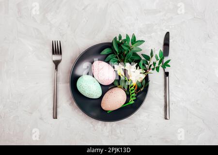 Painted Easter eggs and freesia white flower on black plate on gray textured background. Holiday table setting. Top view, flat lay. Stock Photo