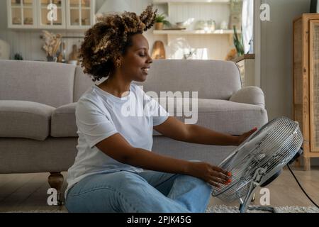Satisfied happy young black woman sitting on floor in front of electrical fan cooling down at home Stock Photo