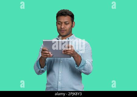 Portrait of a young thoughtful indian guy holding tablet pc. South asian man readin gsomething on his device. Isolated on green background. Stock Photo