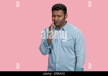 Sad unhealthy indian or arabian guy touching cheek with hand with painful expression. Toothache or dental illness. Isolated on pink. Stock Photo