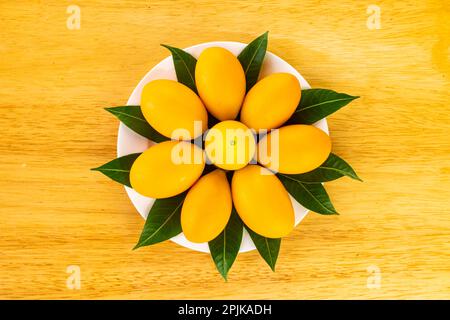 Top view of ripe sweet yellow marian plum or Bouea macrophylla griffith or maprang or mayongchid with green leaves decorated in white ceramic dish on Stock Photo
