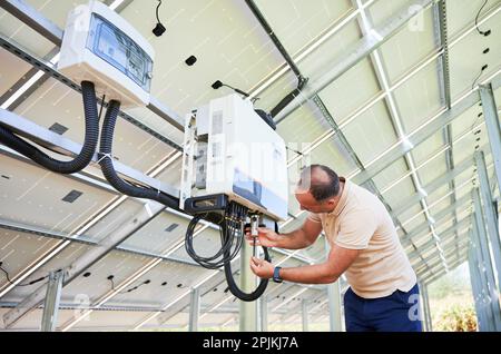 Young man adjusting hybrid inverter. Worker connecting wires to PV battery. Alternative energy in nowadays. Stock Photo