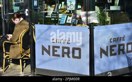 A person sits at a table in the sunshine beside a Caffè Nero sign or notice in Manchester, UK. Stock Photo