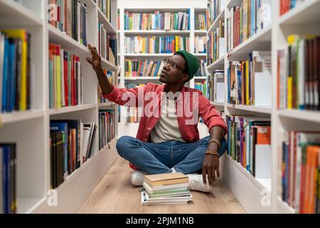 Young African American guy sits on floor in library between bookshelves, student searching for book Stock Photo