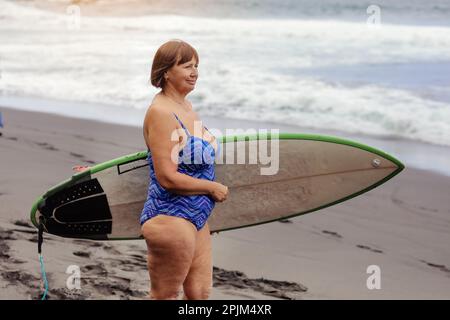 Adult woman in swimsuit holds surfboard to go surfing, enjoys summer vacation at black sand beach by the ocean. Middle aged woman has active lifestyle Stock Photo