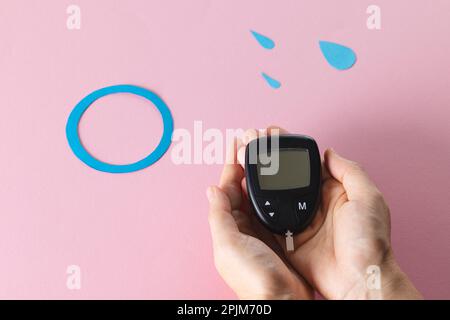 Hands of caucasian woman holding glucometer over blue drops and ring on pink background Stock Photo