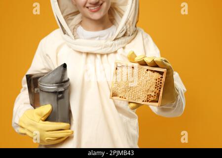 Beekeeper in uniform holding smokepot and hive frame with honeycomb on yellow background, closeup Stock Photo
