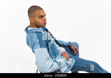 Serious focused young adult Black man in all jeans clothes, sitting alone on plastic chair and posing over white background. High quality photo Stock Photo