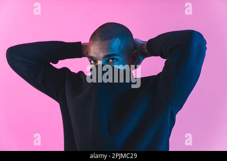 Male collage student posing for fashion photoshoot with hands behind his neck and face covered with hoodie collar. Pink background. High quality photo Stock Photo