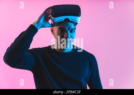 Modern free time activities. Confident young adult man taking off virtual reality headset goggles over pink background. High quality photo Stock Photo