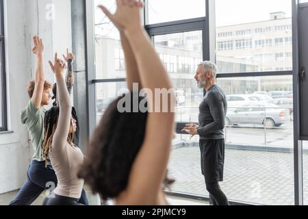 Side view of mature coach talking to multiethnic people standing in Crescent Lunge pose in yoga studio,stock image Stock Photo