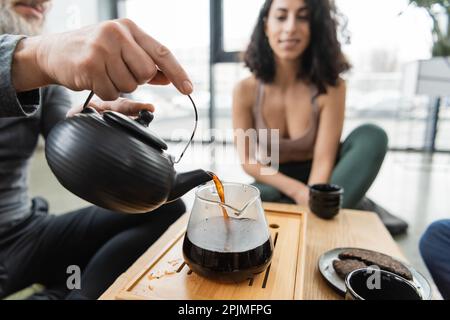 middle aged man pouring brewed puer tea into glass jug near middle eastern woman on blurred background,stock image Stock Photo