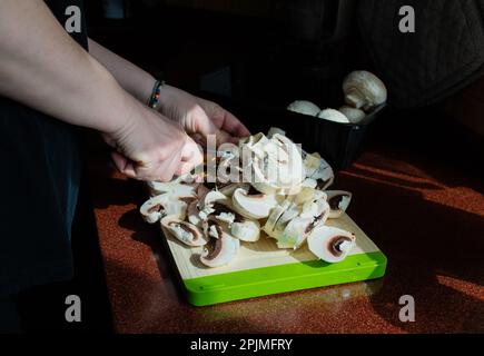 Close-up of hands with sliced fresh mushrooms on a cutting board. Homemade vegetarian cuisine. Sunlight from window, shadows on the table. Stock Photo