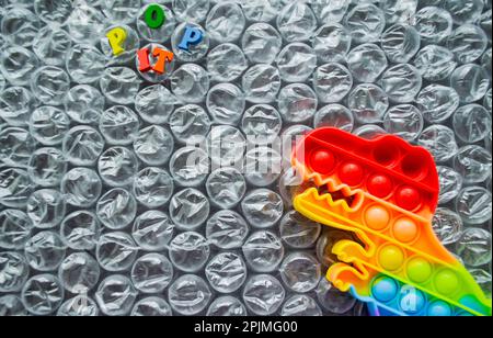 Toy pop it dinosaur rainbow colors on a bubble wrap background with multicolored letters - Pop it. Copy space. Stock Photo