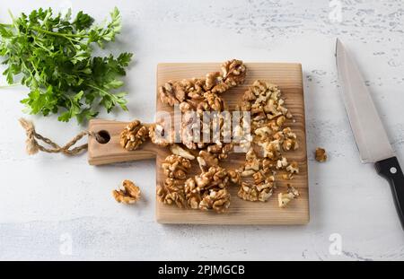 Wooden board of chopped walnuts and greens with a knife on a light gray textured background, home cooking, top view Stock Photo
