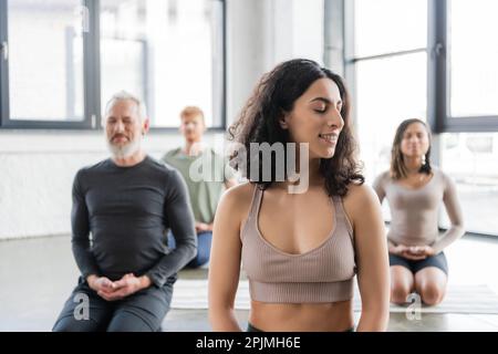 Smiling middle eastern woman meditating near blurred people in yoga class,stock image Stock Photo