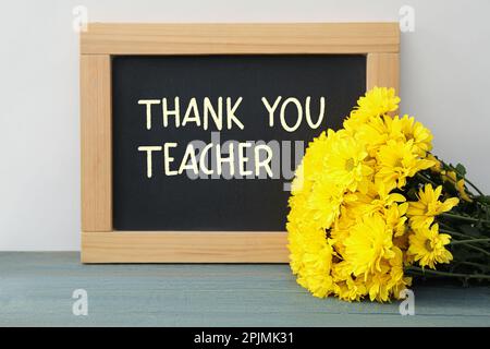 Blackboard with phrase Thank You Teacher and flowers on table near white wall Stock Photo