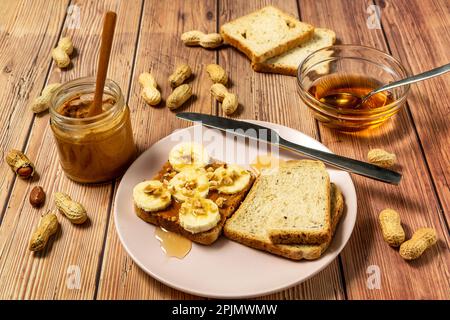 Peanut butter or peanut paste in an open jar and spread on a toast sandwich with banana and honey and peanuts in shells scattered on the table Stock Photo