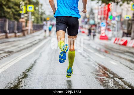 back male athlete in compression socks running marathon in city, summer sports race, jogging on wet asphalt after rain Stock Photo