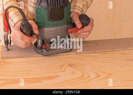 Crop anonymous male carpenter using electric milling machine while cutting wood plank in home workshop Stock Photo