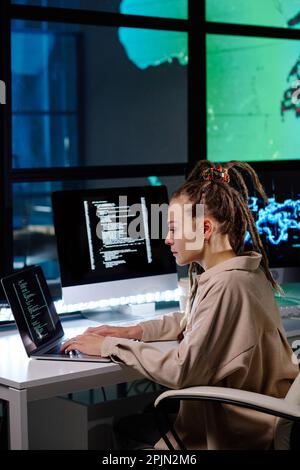 Side view of young female IT support manager with dreadlocks sitting by workplace, looking at laptop screen and typing in dark office Stock Photo