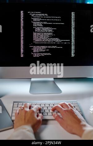 Hands of young female programmer typing on keyboard while sitting by workplace in front of computer screen and decoding data Stock Photo
