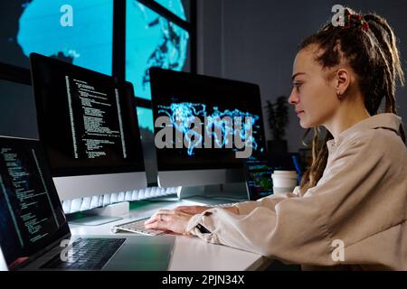 Side view of young programmer developing new software or decoding data on computer screen while sitting by workplace in dark office Stock Photo