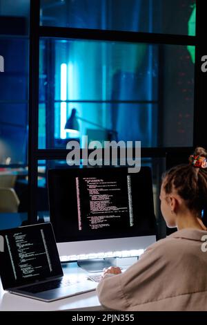 Young female software developer or programmer working with coded data while sitting by workplace in front of computer monitor and laptop Stock Photo