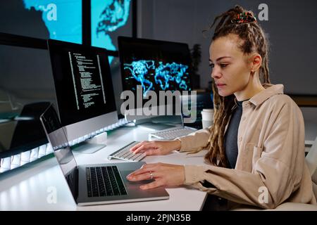 Young serious female programmer looking at data on laptop screen while sitting by workplace in dark office and working overtime Stock Photo