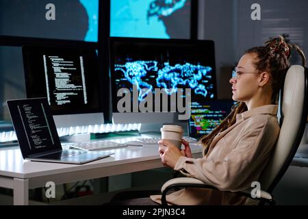 Young serious businesswoman with cup of coffee sitting in armchair by her workplace and looking at computer screen with coded data Stock Photo