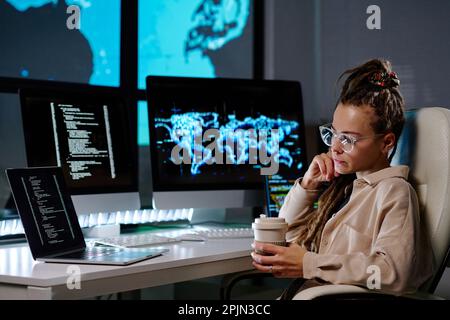 Serious young programmer in eyeglasses looking at coded data on laptop screen and having coffee while sitting by workplace in dark office Stock Photo