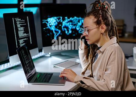 Young busy IT engineer in eyeglasses taking call of client and checking information in laptop while looking through data on screen Stock Photo