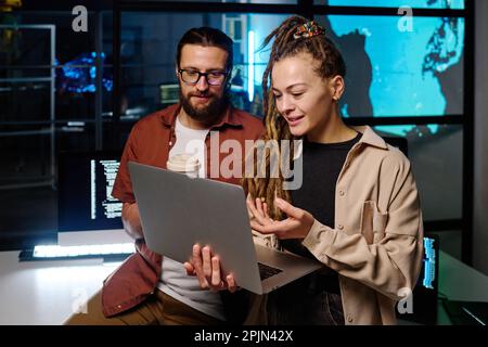 Young confident woman pointing at laptop screen while explaining online data and ways of decoding to male colleague at meeting in office Stock Photo