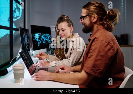 Young confident female programmer pointing at laptop screen with coded data while exlaining something to male colleague at night meeting Stock Photo