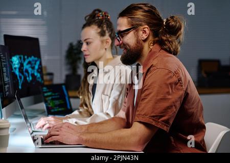 Side view of young male IT engineer typing on laptop keyboard while decoding data on screen against female coworker using computer Stock Photo