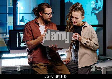 Two young IT engineers discussing online data on laptop screen while man explaining female colleague with cup of coffee simplest way of decoding Stock Photo