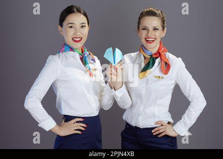 smiling modern flight attendant women in blue skirt, white shirt and scarf with paper airplane against gray background. Stock Photo