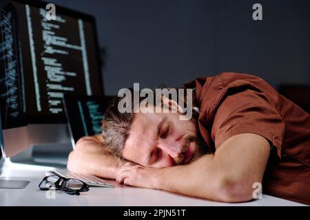 Young exhausted male programmer with his head on desk by computer keyboard sleeping after work over developing new decoding software Stock Photo