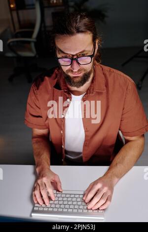 Young male programmer in eyeglasses looking at computer screen and typing on keyboard while sitting by desk and decoding data Stock Photo