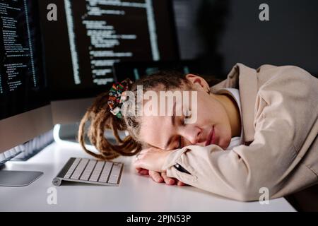 Young exhausted female IT engineer sleeping with her head on desk by computer keyboard while sitting in front of screens with coded data Stock Photo