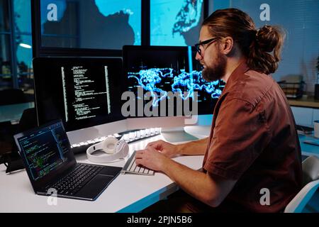 Young serious male programmer looking at coded data on computer screen while sitting by workplace in dark office and working at night Stock Photo