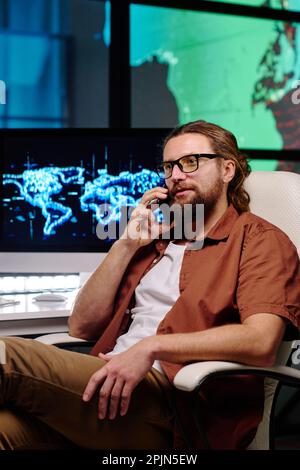 Young confident programmer with smartphone by ear sitting in armchair by workplace with desktop computer and consulting with colleague Stock Photo
