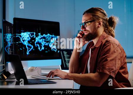 Side view of young busy male IT engineer giving consultation on mobile phone while sitting in front of computer and decoding data Stock Photo
