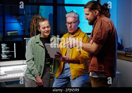Young confident man pointing at tablet screen during presentation of new software to his colleagues at working meeting in dark office Stock Photo