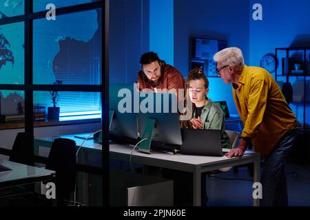 Three colleagues working over new project or software in front of desktop computers while young woman explaining something to men Stock Photo