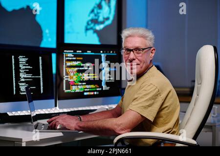 Confident senior male programmer looking at camera while sitting by workplace in dark office at night and using new software Stock Photo